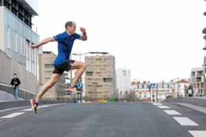 Homme en tee-shirt bleu traversant une route en simulant un saut en course.