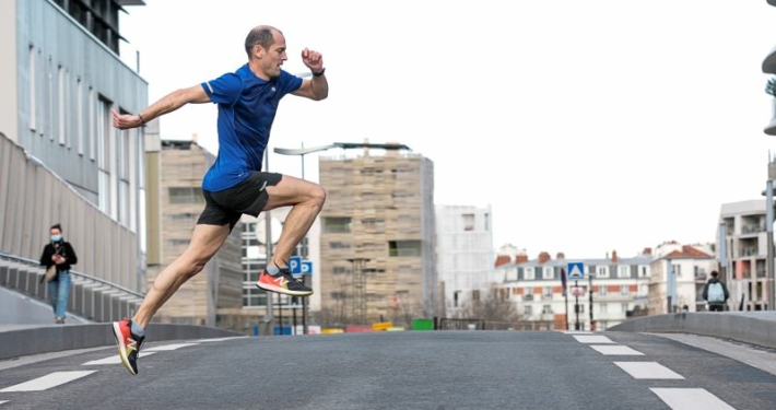Homme en tee-shirt bleu traversant une route en simulant un saut en course.