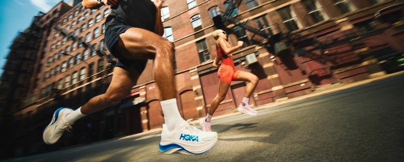 Deux coureurs en mouvement au milieu d'une rue citadine, avec les chaussures HOKA.