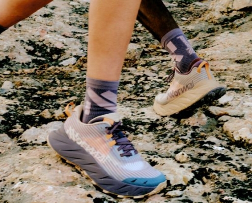 Focus sur la paire de NNormal Cadi, en exposition aux pieds de coureurs sur un sol rocheux