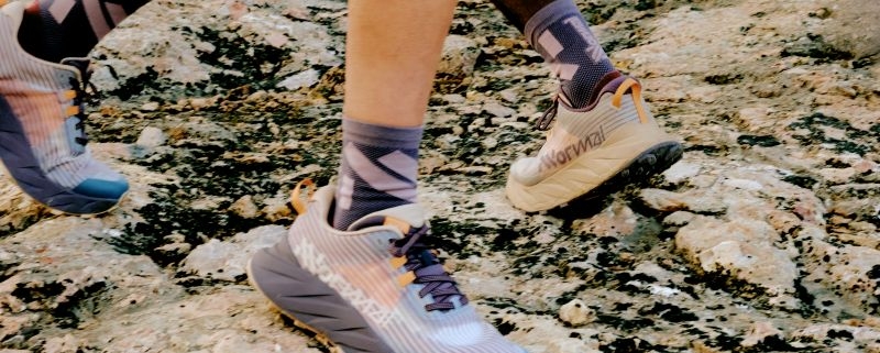 Focus sur la paire de NNormal Cadi, en exposition aux pieds de coureurs sur un sol rocheux